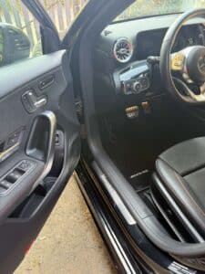 Intérieur noir d'une Mercedes-Benz AMG, porte ouverte, pédales sport, seuil de porte "Mercedes-Benz", commandes de lève-vitre et réglage de siège visibles.