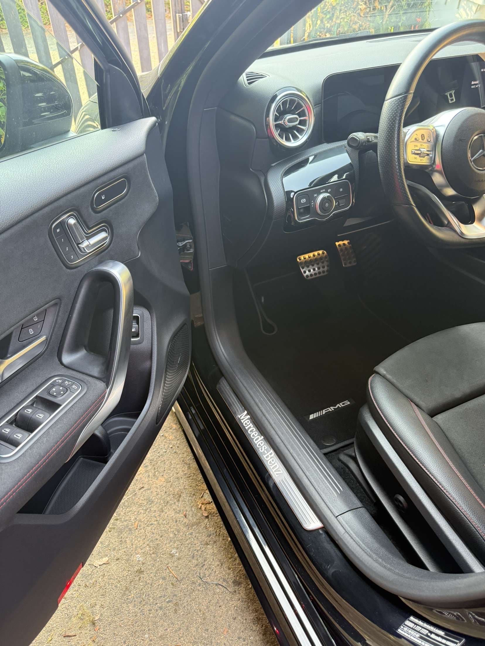 Intérieur noir d'une Mercedes-Benz AMG, porte ouverte, pédales sport, seuil de porte "Mercedes-Benz", commandes de lève-vitre et réglage de siège visibles.