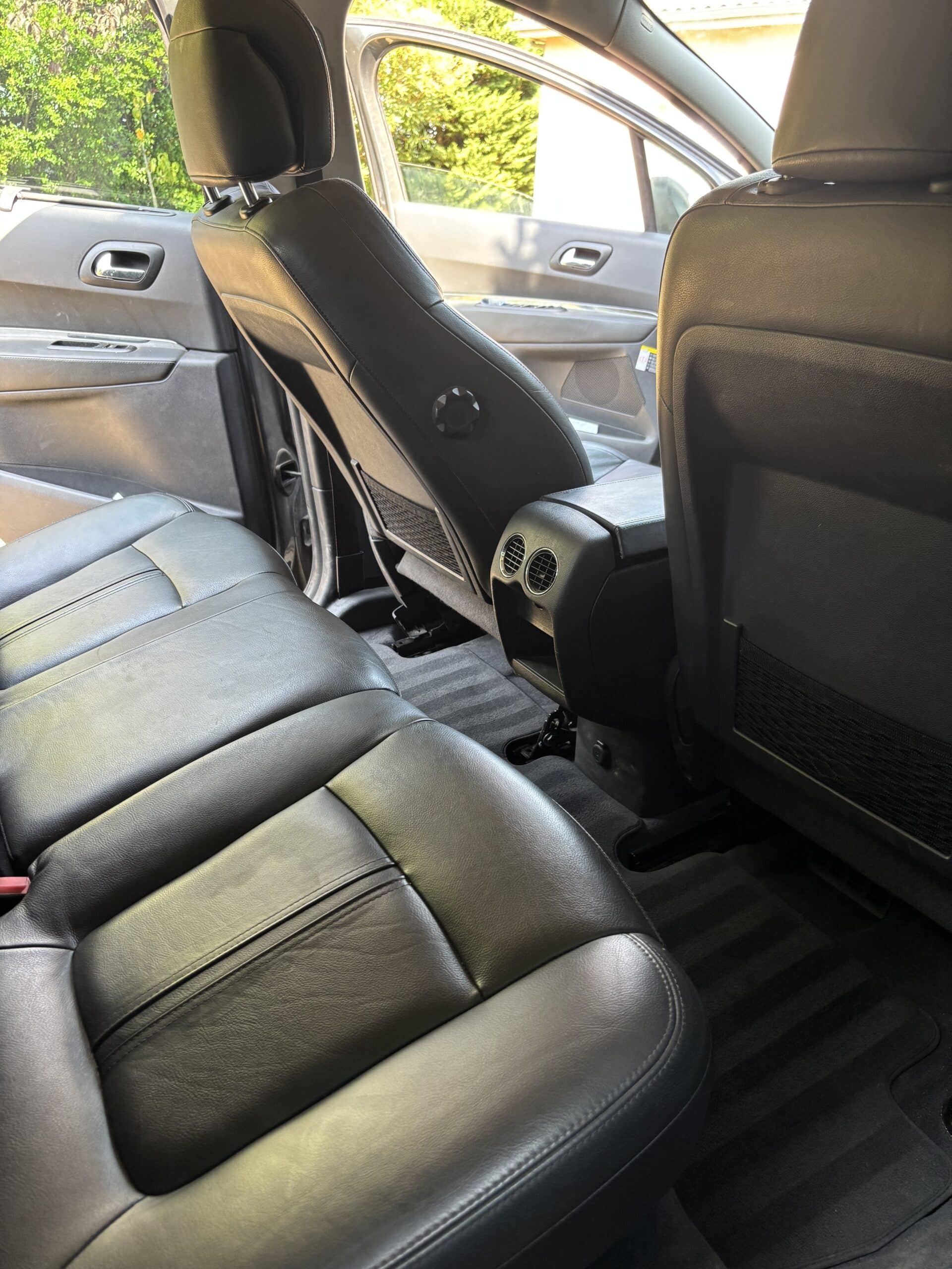 Vue intérieure arrière d'une voiture avec sièges en cuir noir et tapis de sol rayé, éclairée par la lumière extérieure verte.