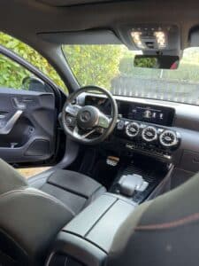 Intérieur Mercedes Classe A, vue du poste de conduite noir avec surpiqûres rouges. Écran MBUX et aérateurs 'turbine'. Porte ouverte.