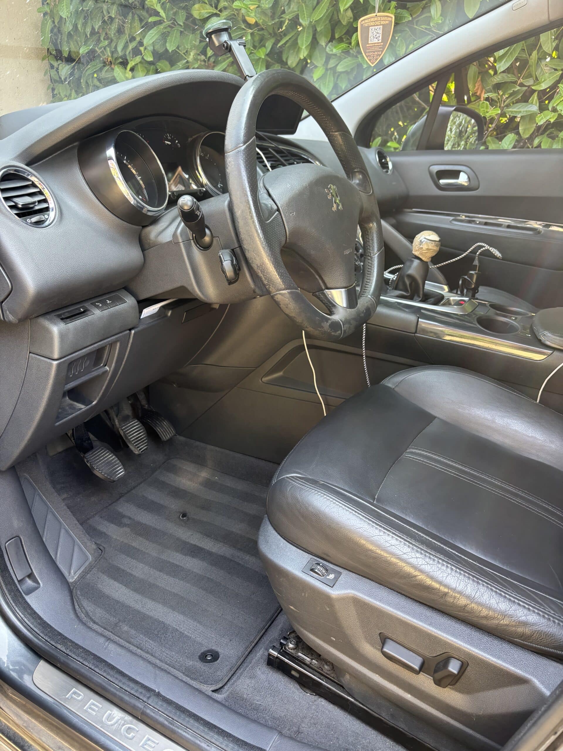 Intérieur avant gauche Peugeot: volant usé, pédalier, siège cuir noir et inscription PEUGEOT sur seuil de porte.