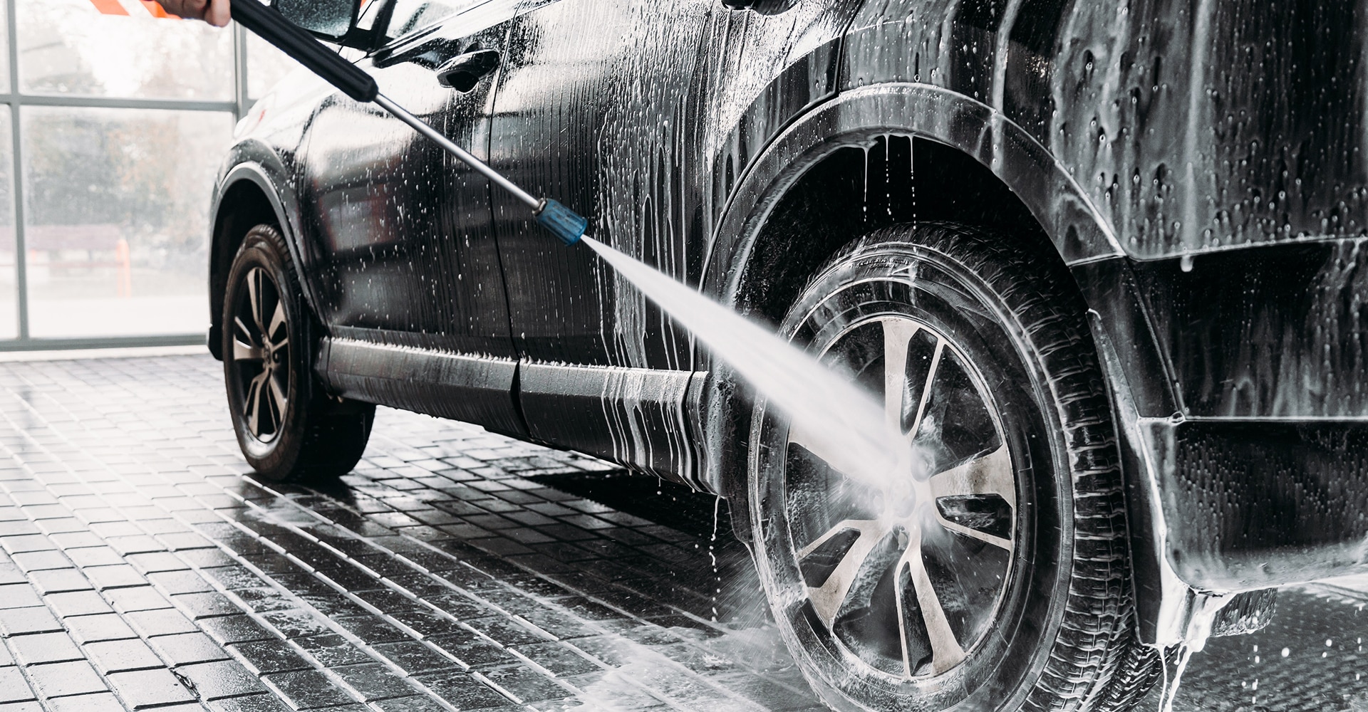 Nettoyage haute pression d'une voiture noire savonneuse, projetant de l'eau sur la jante et le pneu dans une station de lavage.