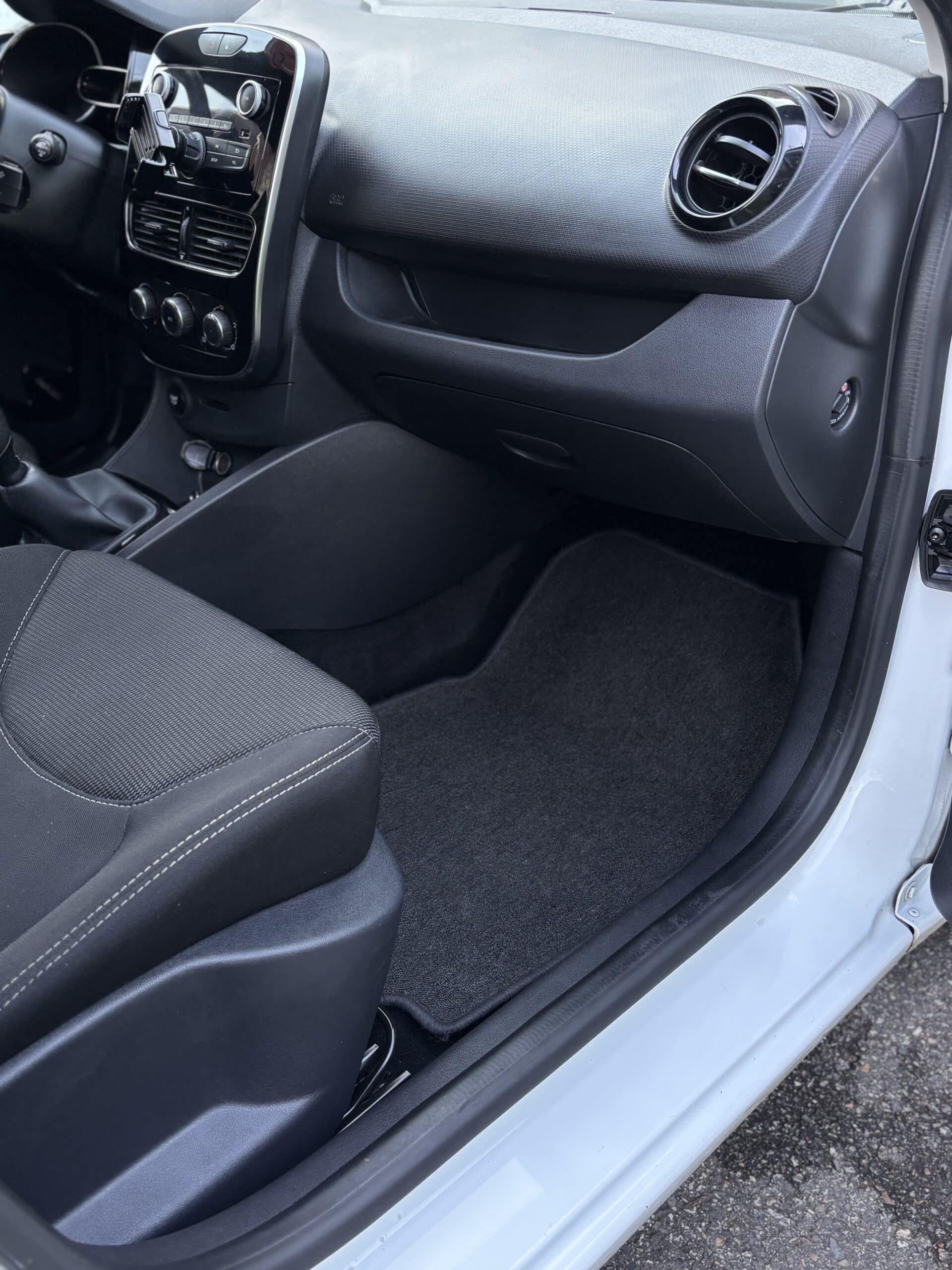 Intérieur d'une Renault Clio avec tableau de bord noir, tapis de sol propre et siège passager en tissu gris.