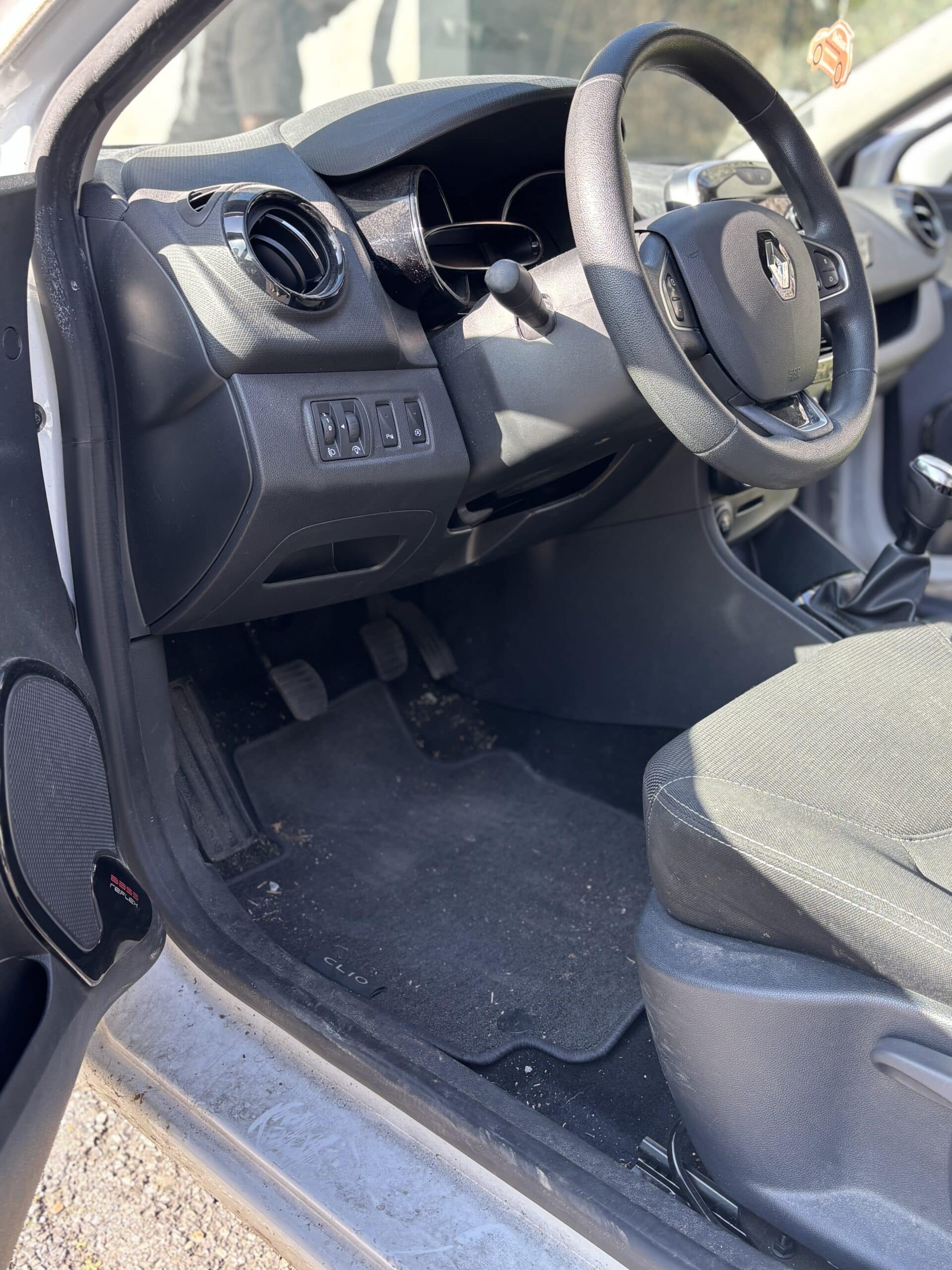 Intérieur d'une Renault Clio montrant le volant, le tableau de bord et les pédales sur un tapis de sol siglé Clio.