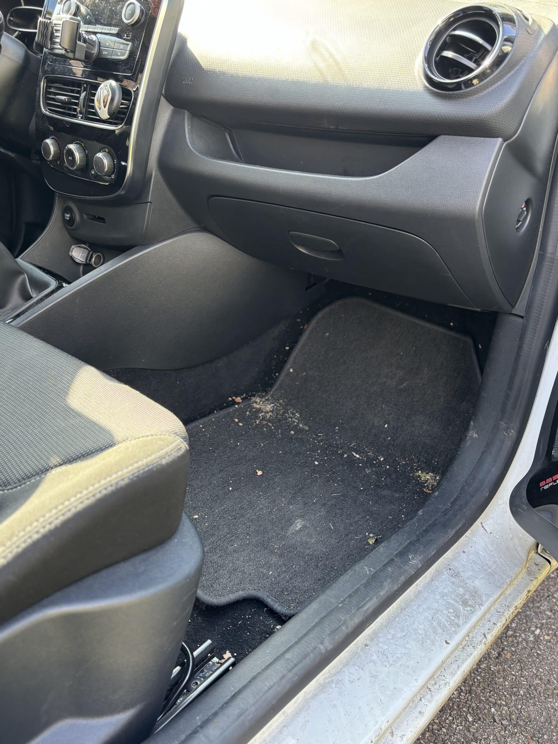 Intérieur de voiture avec tapis de sol sale, miettes et débris côté passager près du tableau de bord noir.